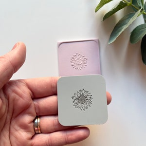 May include: A close-up of a hand holding two silicone stamps, one with a raised sunflower design and the other with a flat sunflower design.