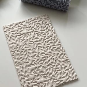 May include: A light gray rolling pin and a rectangular clay slab, both with a repeating leaf pattern. The rolling pin is cylindrical, and the slab is rectangular. The leaf pattern is raised on both items, creating a textured surface.