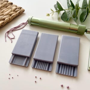 Peut inclure: Ensemble d'outils artisanaux, dont trois outils rectangulaires gris avec des bords striés, une poignée en métal vert et de petites perles roses. Un tapis tissé et de la verdure sont également visibles.