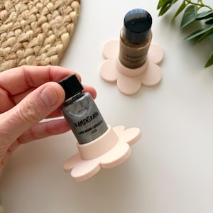 May include: A bottle of silver liquid cosmetic product is held in a light pink flower-shaped holder. The product is labeled "KAMENSKAYA SUPER SHINING LIQUID GLITTER SILVER". Another bottle sits on a similar holder.