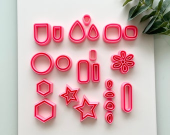 May include: A collection of pink polymer clay cutters in various shapes, including geometric forms, stars, and a flower. The cutters are arranged on a white surface, perfect for crafting earrings or other jewelry.