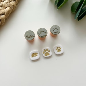 Peut inclure: Ensemble de tampons avec des motifs d'ours et d'empreintes de pattes. Les tampons ont un motif gris sur une base pêche clair. Les tampons carrés ont une base blanche avec des motifs d'ours et d'empreintes de pattes dorés. Les tampons sont disposés sur une surface blanche.