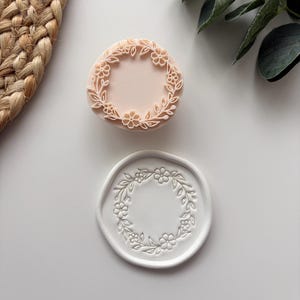 May include: A round, light peach-coloured stamp featuring a raised floral wreath design and a corresponding white circular wax seal impression showing the same intricate botanical pattern of small flowers and leaves in a delicate, embossed relief.