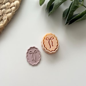 Könnte beinhalten: Zwei ovale Ausstecher mit einem Schleifendesign. Einer ist hellrosa, der andere pfirsichfarben. Die Ausstecher haben einen gewellten Rand und sind aus Kunststoff. Der Hintergrund ist weiß mit einem geflochtenen Korb und Grünzeug.