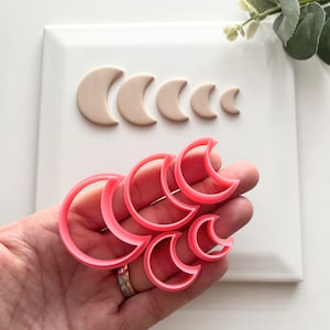 May include: A selection of pink crescent moon-shaped cookie cutters in different sizes, held in a hand. Several beige crescent moon shapes are arranged on a white surface. The cutters are made of plastic and are used for baking or crafting.