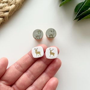 May include: A collection of four decorative earrings. Two are round, gray, and feature a silver deer design. The other two are square, white, and have a gold deer design. The earrings are small and appear to be handmade.