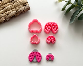 May include: A set of pink plastic cookie cutters in the shape of ladybugs. The cutters are in different sizes and have a detailed design. There are also two pink ladybug-shaped cookies.