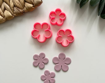 Puede incluir: Cortadores de galletas rosas con forma de flor y recortes de flores lavanda dispuestos sobre una superficie blanca. Los cortadores son de plástico, y los recortes parecen ser de cuero. Una cesta tejida y vegetación se ven en el fondo.