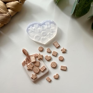 May include: A heart-shaped silicone mould filled with various shapes, including stars, hearts, and rectangles. A small cutting board with biscuit-shaped resin pieces is also present, along with loose biscuits. The biscuits are light brown.