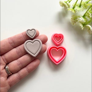 May include: Set of three heart-shaped cookie cutters. The cutters are nested, with the largest cutter being pink and the two smaller cutters being a light grey colour.