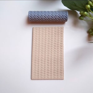 May include: A gray rolling pin with a repeating pattern of wavy lines. The rolling pin is on a white surface next to a beige piece of clay with the same pattern embossed into it.