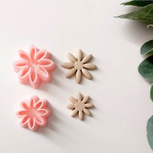 Könnte beinhalten: Vier blütenförmige Ausstecher, zwei rosa und zwei beige. Die rosa Ausstecher sind übereinander gelegt, während die beige Ausstecher einlagig sind. Die Ausstecher befinden sich auf einer weißen Oberfläche.