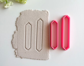 Puede incluir: Dos cortadores de galletas de plástico rosa en forma de rectángulos largos y estrechos con bordes redondeados. Los cortadores están sobre una superficie blanca con un trozo de arcilla gris detrás. La arcilla se ha presionado con los cortadores para mostrar la forma.