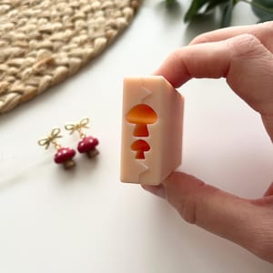Puede incluir: Un molde rectangular rosa claro con tres recortes de hongos. Se ven un par de pendientes de hongo rojos con lazos dorados. Los artículos están sobre una superficie blanca con una estera tejida en el fondo.