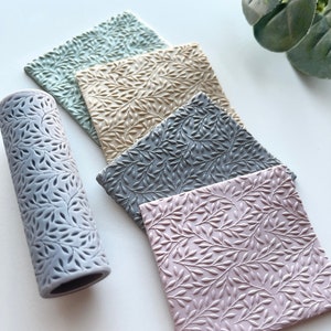 Puede incluir: Cuatro tapetes cuadrados de silicona con un patrón de hojas en diferentes colores: azul claro, beige, gris y rosa. También se muestra un rodillo de silicona gris con el mismo patrón de hojas.