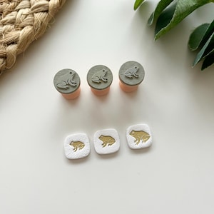 May include: A collection of frog-themed stamps and stamp pads. The stamps feature a frog design on a gray circular base, atop a peach-colored cylindrical pad. Three square stamps with gold frog designs are also present.