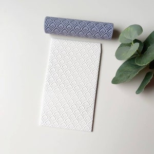 May include: A gray rolling pin with a geometric pattern, used to create an embossed design on a white surface. The surface has a repeating pattern of small squares.
