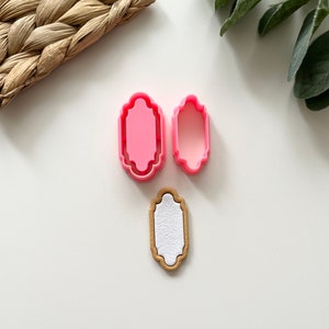 以下が含まれることがあります： 丸みを帯びた角と装飾的な縁取りのある長方形のピンクのプラスチック製のクッキーカッターが2つ。白い中心を持つ茶色のクッキーも表示されています。