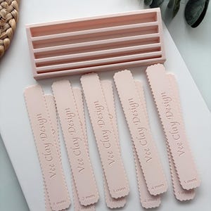 May include: A light pink rectangular organiser tray with five horizontal slots and a set of ten light pink scalloped edge clay thickness guides. Each guide is embossed with the text Vee Clay Design and specific measurements from 1 mm to 5 mm.