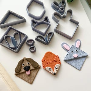 May include: A collection of grey plastic cookie cutters in diverse shapes, such as a triangle, hexagon, and animal faces. Also, three handmade leather bookmarks shaped like a dog, fox, and rabbit, with detailed features.