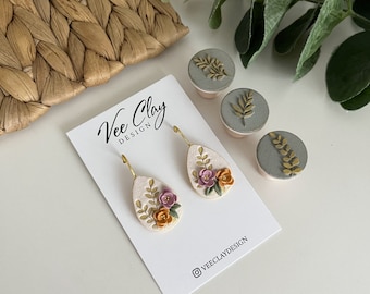 May include: A pair of dangle earrings with a white teardrop-shaped base and gold accents. The earrings feature a floral design with pink, orange, and yellow flowers and green leaves. The earrings are displayed on a white card with the text "Vee Clay Design" and the Instagram handle "@veeclaydesign".