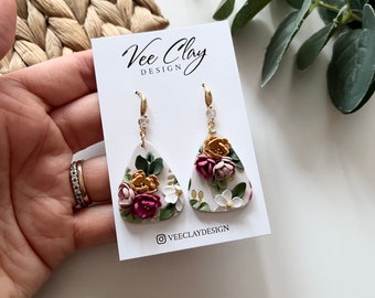 May include: A pair of white triangle-shaped earrings with a floral design. The earrings feature pink, white, and yellow flowers with gold accents. The earrings are on a white card with the text "Vee Clay Design" and the Instagram handle "@veeclaydesign".