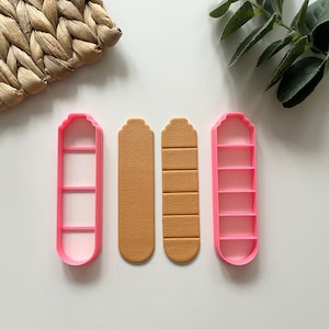 Op de afbeelding: Twee roze plastic koekjesvormen in de vorm van lange rechthoeken met afgeronde uiteinden en vijf horizontale lijnen uitgesneden. Twee bruine fondantstukken zijn in het midden te zien, uitgesneden met de koekjesvormen.