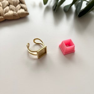 Könnte beinhalten: Ein goldfarbener, verstellbarer Ring mit rechteckiger Fassung, neben einer kleinen rosa quadratischen Form. Der Ring hat ein Doppelband-Design. Die Artikel befinden sich auf einer weißen Oberfläche, mit grünem Laub und einem gewebten beigen Artikel im Hintergrund.