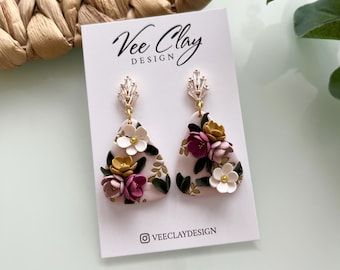 May include: A pair of pink and white floral dangle earrings with gold accents. The earrings are made of polymer clay and feature a variety of flowers, including pink, white, and yellow. The earrings are attached to gold hooks.