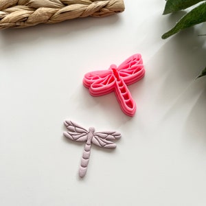 May include: Two decorative dragonfly shapes, one pink and one light purple, on a white background.