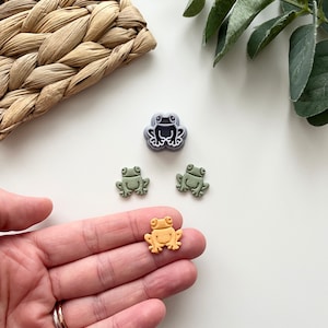Puede incluir: Tres cortadores de galletas con forma de rana, dos verdes y uno naranja, con un cortador de galletas con forma de rana morado en el fondo.