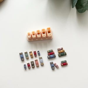 Pode incluir: Uma coleção de contas de argila polimérica em miniatura em forma de livro, em várias cores, incluindo azul, vermelho e dourado. Um molde rosa claro com cavidades em forma de livro também está presente. Ideal para artesanato.