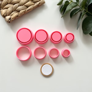 May include: A set of eight pink plastic circle cookie cutters of varying sizes, with a single wooden circle with a white center.