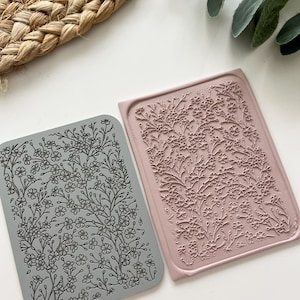 May include: Two rectangular floral design stamps. One stamp is gray, and the other is a dusty rose color. The stamps feature a detailed pattern of flowers and leaves. The stamps are likely used for crafting or scrapbooking projects.