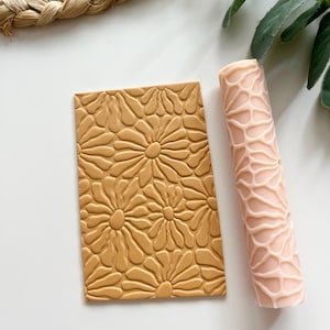 Puede incluir: Un sello rectangular de arcilla color beige con un patrón de margaritas y un rodillo rosa con un diseño texturizado. Estos artículos son probablemente para manualidades o repostería, ofreciendo un elemento decorativo.