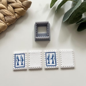 May include: A gray plastic mold with the word "FISH" and four white rectangular stamps. Two stamps feature a blue fish design. The stamps have a dotted border. The mold is square and has a grid-like pattern.