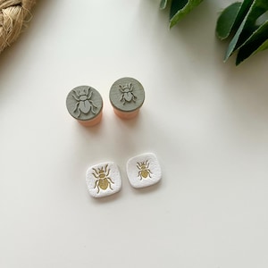 May include: Two round, grey wooden stamps with insect designs, atop peach-coloured bases, and two white square earrings with gold insect designs. The items are arranged on a white surface, with a woven texture and green leaves in the background.