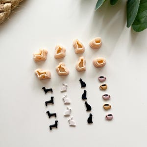 May include: A collection of miniature polymer clay charms in various shapes. Includes dachshunds, rabbits, cats, and coffee cups in black, white, and tan colours. Also, there are several tan-coloured, abstract shapes. The charms are arranged on a white surface.