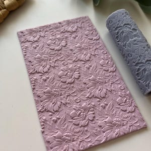 Puede incluir: Un rodillo de silicona de color lila claro con un diseño floral. El rodillo se utiliza para crear patrones en relieve en fondant o arcilla.