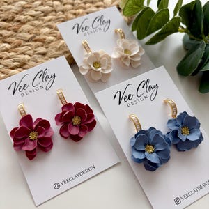 Puede incluir: Tres pares de pendientes florales en crema, burdeos y azul, cada uno con un aro dorado y un pequeño centro dorado. Los pendientes se exhiben en tarjetas blancas con el texto "Vee Clay Design" y un identificador de redes sociales.