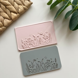 Może przedstawiać: Dwa silikonowe stemple z kwiatowym wzorem. Górny stempel jest różowy, a dolny szary. Oba stemple przedstawiają wzór kwiatów, liści i motyli.