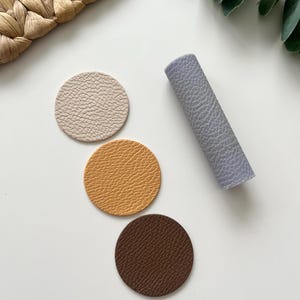 Könnte beinhalten: Drei runde Lederuntersetzer in Beige, Orange und Braun. Eine Rolle aus helllila strukturiertem Leder ist ebenfalls abgebildet.