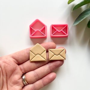 以下が含まれることがあります： ピンク色の封筒型クッキーカッター2つと、ベージュ色の封筒型クッキー2つ。クッキーカッターはプラスチック製で、開いた封筒と閉じた封筒の形をしています。クッキーはベージュ色で、表面にテクスチャがあります。