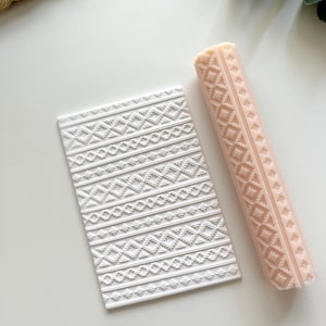 May include: A white rectangular textured stamp and a light pink rolling pin with a geometric pattern. The stamp and rolling pin are likely for baking or crafting, featuring a repeating diamond and line design.