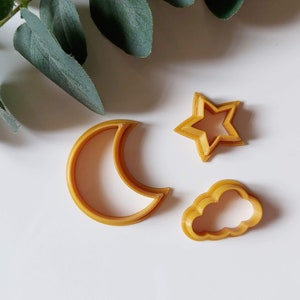 Puede incluir: Tres cortadores de galletas de color dorado: una luna creciente, una estrella y una nube.