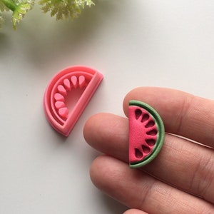 Pode incluir: Um amuleto de argila polimérica em forma de fatia de melancia rosa e verde. O amuleto é feito para parecer uma fatia de melancia com casca verde e polpa vermelha. O amuleto tem aproximadamente 2,5 cm de diâmetro.