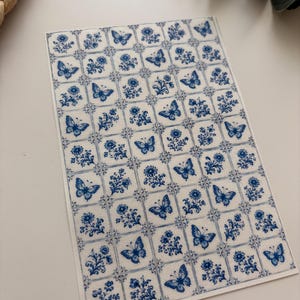 Peut inclure: Une feuille rectangulaire d'autocollants présentant un motif en grille de carrés individuels. Chaque carré contient une illustration à l'encre bleue d'un papillon ou d'un brin floral sur fond blanc, créant un design répétitif en damier.