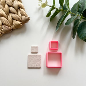 May include: A square-shaped earring and three pink square-shaped polymer clay cutters. The earring is a light beige color with a textured surface. The cutters are in three different sizes. A woven straw placemat and green leafy sprig are also in the image.