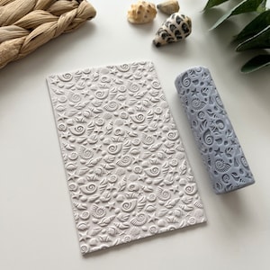 Puede incluir: Un rodillo gris con un patrón de conchas marinas y una hoja texturizada blanca con un patrón de conchas marinas.