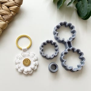 May include: A white flower-shaped keychain with a gold ring and a small gold circle with the letter "V" in the center. The keychain is next to three gray plastic flower-shaped cookie cutters.